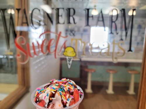 A close-up of a dessert cup topped with ice cream and colorful sprinkles, with a sign for Wagner Farm & Sweets Treats in the background.