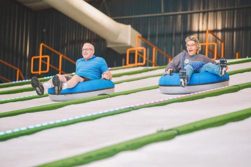 Two people joyfully sliding down a snowy slope on blue inflatable tubes, with colorful lights in the background.