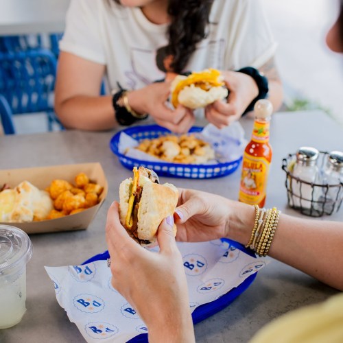 A person holds a sandwich while another enjoys food in the background, with drinks and condiments on the table.