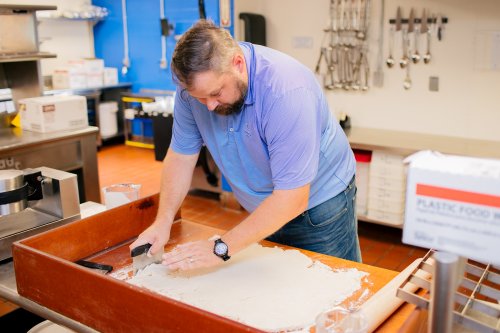 A man in a blue shirt rolls out dough on a wooden surface in a kitchen filled with cooking tools.