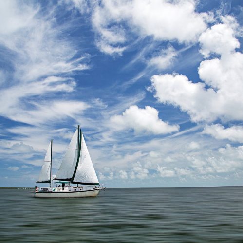 A sailboat glides across calm waters under a bright blue sky with fluffy white clouds.