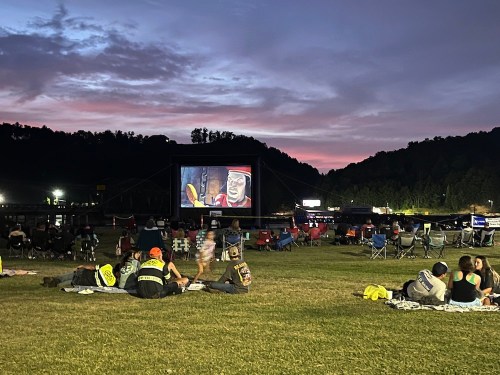 Outdoor movie screening at dusk, with people sitting on the grass and a large screen displaying a film.