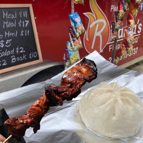 A skewer of grilled meat next to a large steamed bun, with a food truck in the background displaying menu prices.
