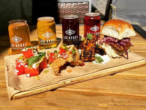 A wooden platter with a burger, grilled chicken, watermelon salad, and colorful drinks in jars.