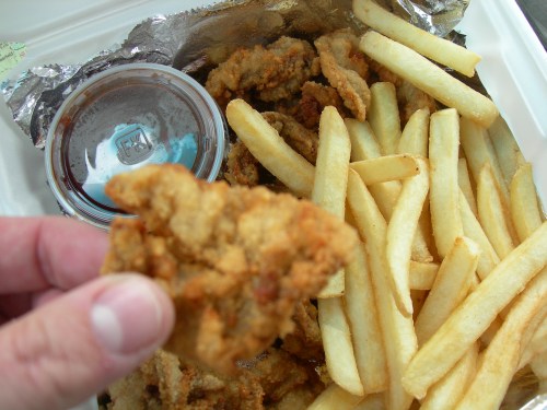 A hand holding a piece of fried chicken above a container of fries and dipping sauce.