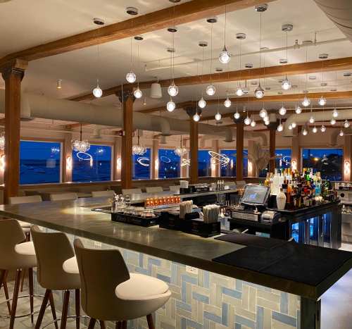 A modern bar with pendant lights, a sleek counter, and a view of the ocean at dusk.