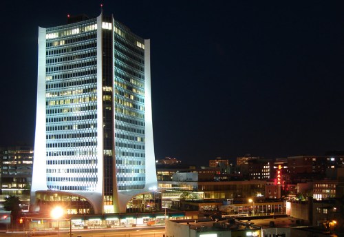 A tall, modern skyscraper illuminated at night, surrounded by city lights and buildings.
