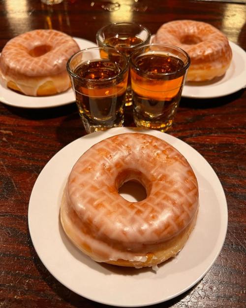 Three glazed donuts on white plates with two shot glasses of amber liquid in the background.