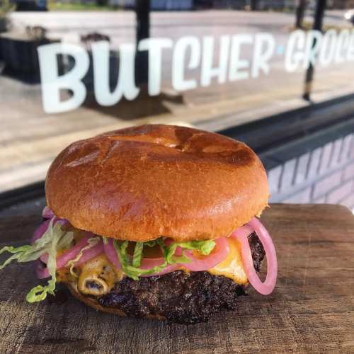 A juicy burger with melted cheese, lettuce, and pickled onions on a toasted bun, set against a shop window.