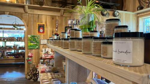 A wooden shelf displaying various scented candles in jars, with a cozy shop interior in the background.