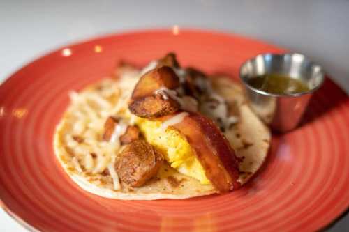 A close-up of a breakfast taco with eggs, bacon, potatoes, cheese, and a small cup of salsa on a red plate.