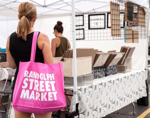 A woman with a pink tote bag stands at a market stall displaying various items under a white tent.