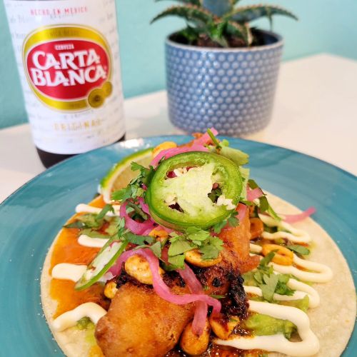 A colorful taco topped with jalapeños, onions, and cilantro, served with a beer and a small plant in the background.
