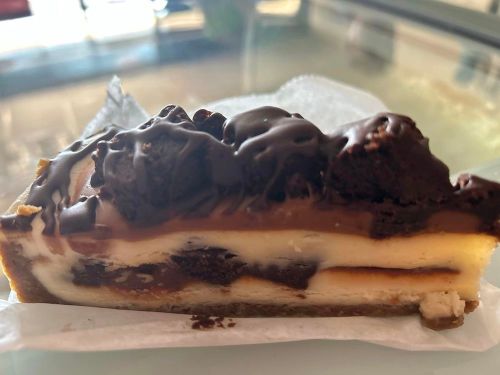 A slice of layered dessert with chocolate topping, brownie pieces, and a creamy filling on a graham cracker crust.
