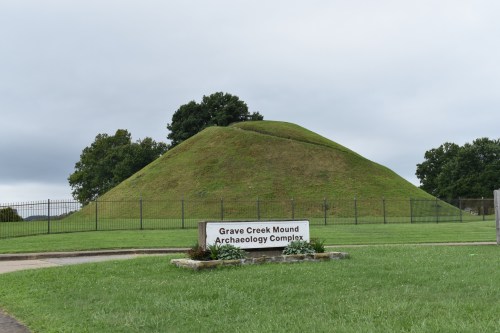 Grave Creek Mound, a large grassy burial mound, with a sign in front, set against a cloudy sky.