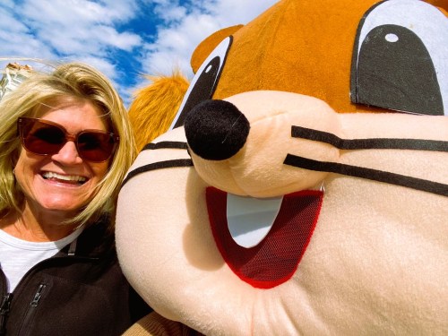 A smiling woman poses with a large, friendly cartoonish animal mascot against a blue sky.