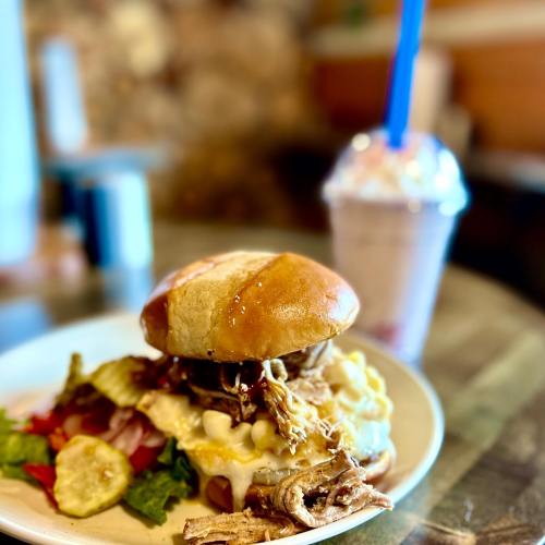 A close-up of a pulled pork sandwich with toppings on a plate, alongside a milkshake in the background.