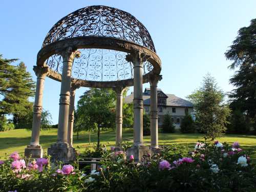 A decorative gazebo surrounded by colorful flowers, with a large house and trees in the background under a clear blue sky.