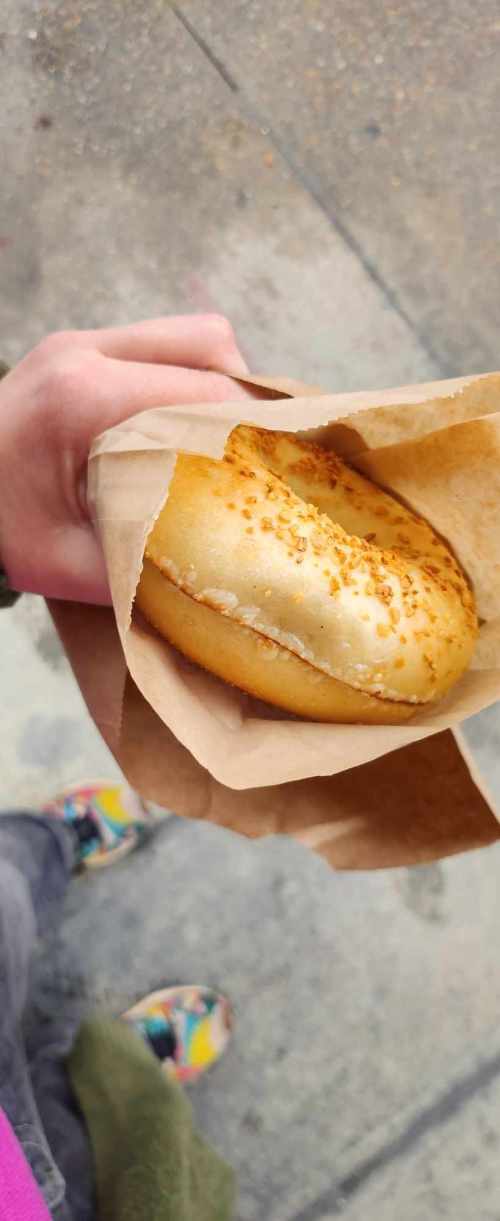 A hand holding a sesame seed bun in a brown paper bag, with colorful shoes visible in the background.
