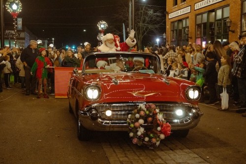 A festive red convertible with Santa and a passenger waves to a crowd during a holiday parade.
