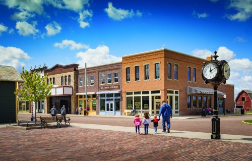 A family walks down a brick street lined with historic buildings and a clock, under a bright blue sky with clouds.