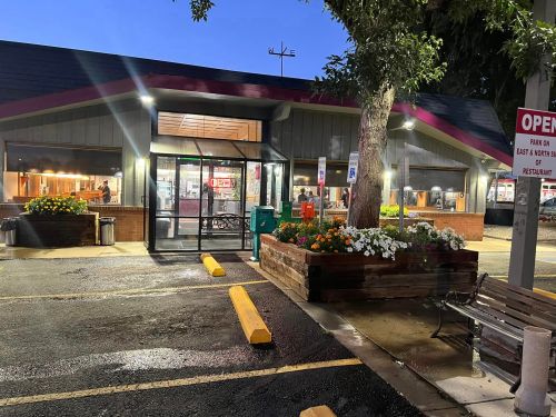 A restaurant exterior at dusk, featuring an "Open" sign, flower beds, and a bench in the foreground.