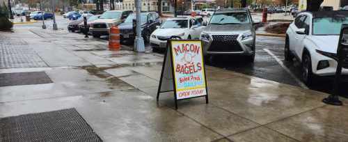 A sidewalk sign for Macon Bagels, advertising fresh bagels and stating "Open Today," with wet pavement and parked cars nearby.