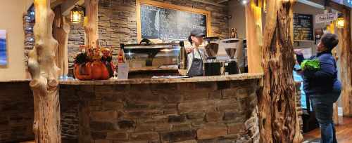 A cozy café with a stone counter, a barista serving a customer, and autumn decorations on display.