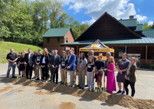 A group of people in formal attire and West Liberty University shirts gather for a groundbreaking ceremony outdoors.