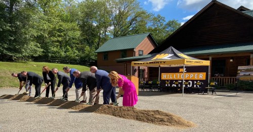 A group of people in business attire participate in a groundbreaking ceremony outdoors, with a tent and a building in the background.
