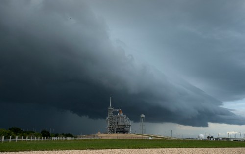 A launch pad under dark, ominous storm clouds, with a grassy foreground and distant structures.