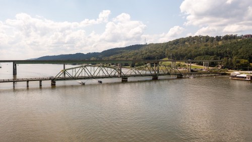 A bridge spans a river, surrounded by green hills and a cloudy sky, with boats visible on the water.