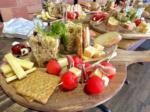 A wooden platter with assorted appetizers, including pasta salad, cheese, crackers, meats, and watermelon.