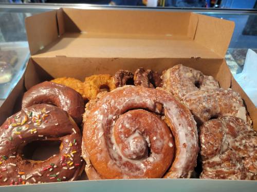 A box of assorted donuts, including chocolate, glazed, and a cinnamon roll, with colorful sprinkles on some.