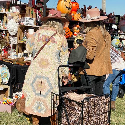 Two women in hats browse a vibrant market filled with decorations and crafts, one wearing a colorful patterned coat.