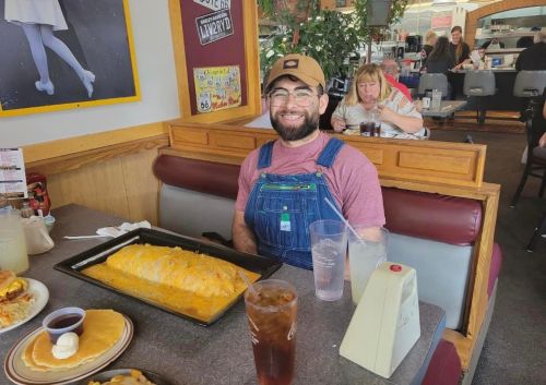 A smiling person in overalls sits at a restaurant table with a large omelet and drinks in front of them.