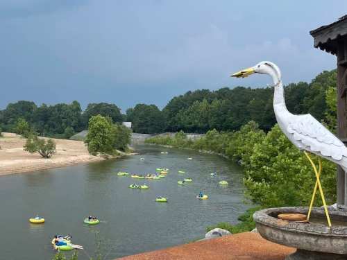 A decorative white heron overlooks a river with people floating on green inner tubes under a cloudy sky.