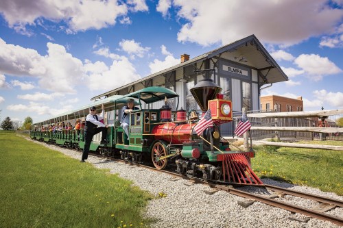 A colorful steam train with passengers at a historic station, surrounded by green grass and a blue sky with clouds.