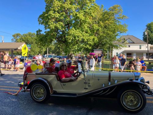 A vintage car with passengers in red shirts drives past a crowd during a sunny parade. Trees and buildings in the background.