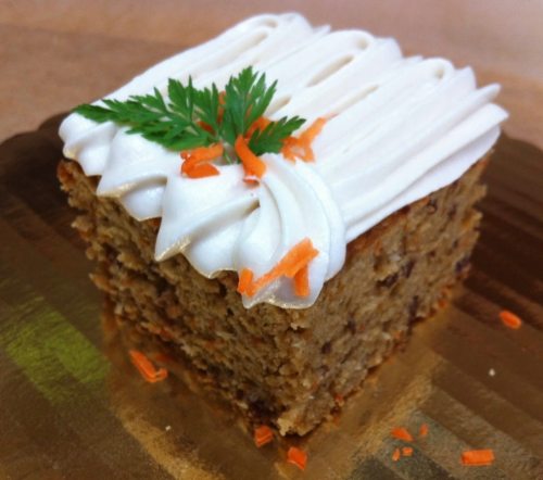 A slice of carrot cake topped with cream cheese frosting and garnished with orange zest and a sprig of parsley.