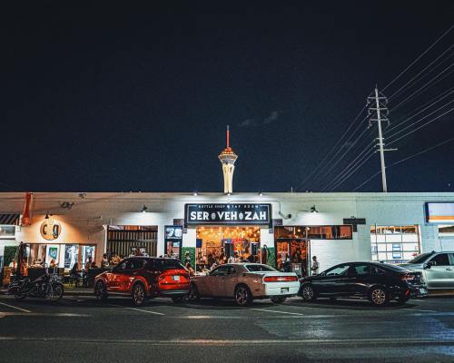 A lively restaurant at night with outdoor seating, featuring a neon sign and the Las Vegas Stratosphere in the background.