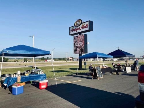 Outdoor market scene with tents, food stands, and a sign for "Wood Bee Antique Mall" under a clear blue sky.