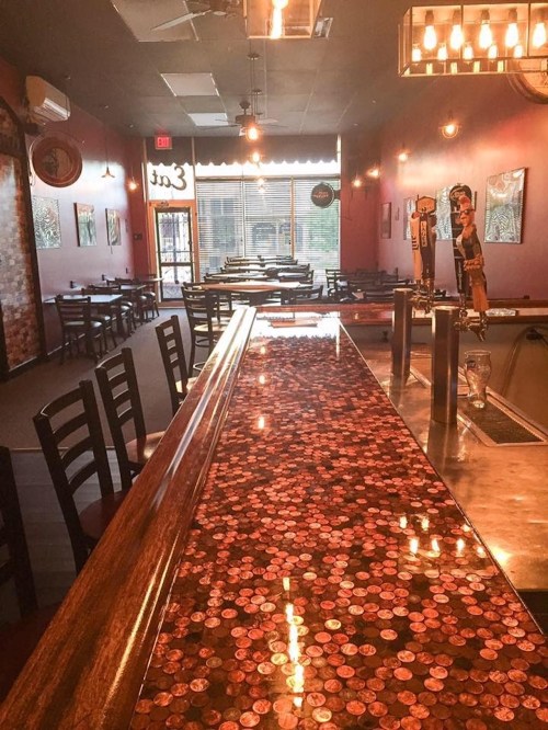 A cozy restaurant interior featuring a bar with a penny-covered countertop and empty tables in warm lighting.