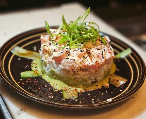 A beautifully plated sushi dish with rice, fish, green onions, and sesame seeds on a dark plate.