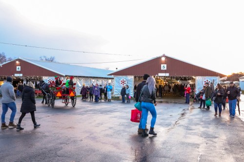People walking outside two large tents at a festive event, with a horse-drawn carriage and holiday decorations.