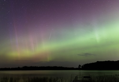 A stunning night sky with vibrant green and purple auroras over a calm lake, dotted with stars and a faint meteor streak.