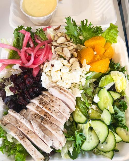 A colorful salad with sliced chicken, mixed greens, cucumbers, almonds, feta, cranberries, pickled onions, and orange segments.