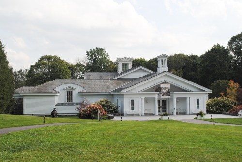 A white building with a large entrance, surrounded by green grass and trees under a cloudy sky.