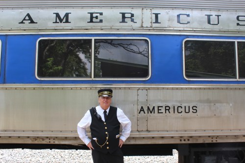 A train conductor stands proudly in front of a vintage train car labeled "Americus."