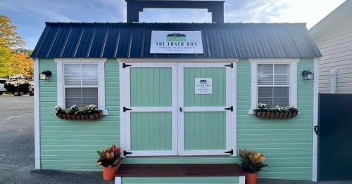 A small, mint green food stand with a sign reading "The Lunch Box" and decorative plants at the entrance.